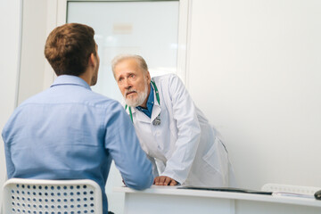 Obraz premium View from back to mature male doctor otolaryngologist performing examination of young man patient throat in office of medical clinic. Sick young man on consultation with sore throat and glands.