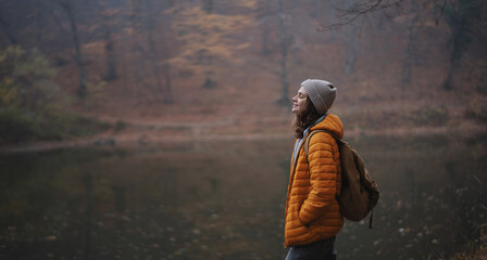 Young woman traveler in a yellow jacket and hat with a backpack breathing fresh air in the autumn...