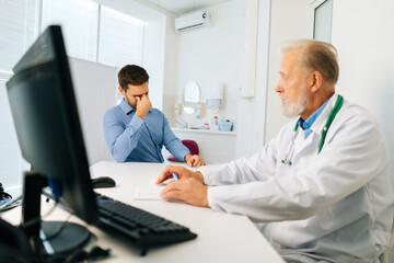 Mature adult male doctor explaining problem to crying sad young man patient sitting at desk during...
