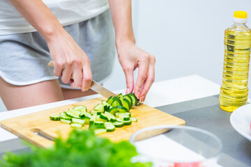 Obraz na płótnie Canvas Close up of a girl hands slicing cucumber with a knife on a cutting board for a vegan vitamin vegetable salad and putting them in a glass bowl while cooking breakfast in the kitchen
