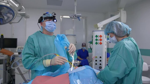 Operating surgeon wearing special equipment on his head in the surgery room. Doctor observes the operation running on the screen. Surgeon applies medical tools to the patient.
