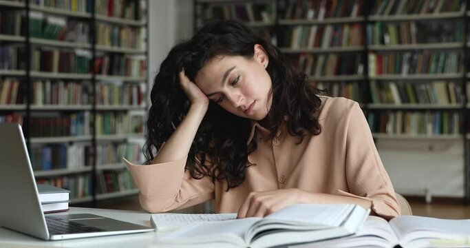 Young tired unmotivated girl student sit at table in library prepares for exam test, feeling lack of ideas while makes coursework. Boring study, monotonous learning, unloving subject at school concept