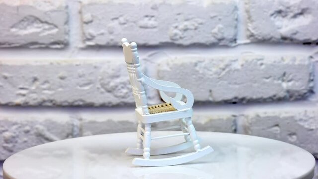 Little white rocking chair for doll’s house. Pretty small piece of furniture on the rotating table. White brickwall at the background. Close up.