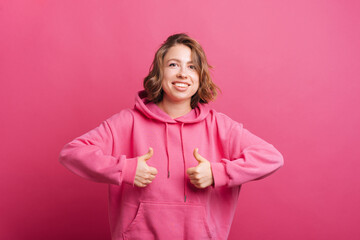 Portrait of young trendy woman in pink hoodie showing thumbs up and smiling at camera.