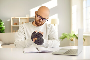 Unhappy man in glasses sitting at desk and looking at his aching hands in compression gloves. Man suffering from acute rheumatoid arthritis pain wears special fingerless gloves for working on computer - obrazy, fototapety, plakaty