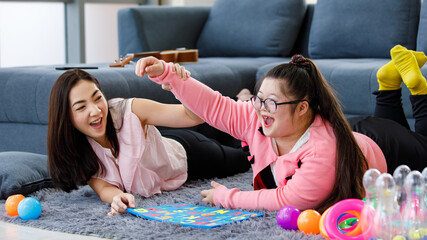 Asian young happy mother laying down on carpet floor near a girl with down syndrome daughter wearing glasses playing smiling laughing and using alphabets puzzle toy teaching together
