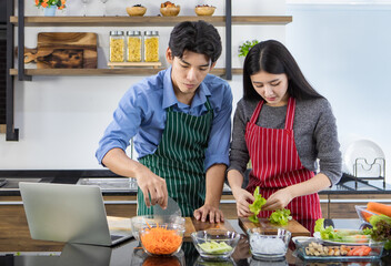 Young Asian chef couple helping each other to cook healthy food of salad from tasty organic vegetable on glass dish by learning recipe instruction from online diet course on laptop