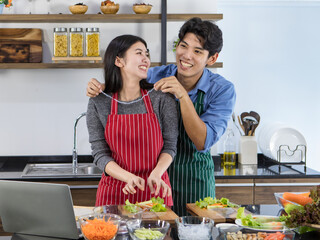 Young Asian chef couple helping each other to cook healthy food of salad from tasty organic vegetable on glass dish by learning recipe instruction from online diet course on laptop