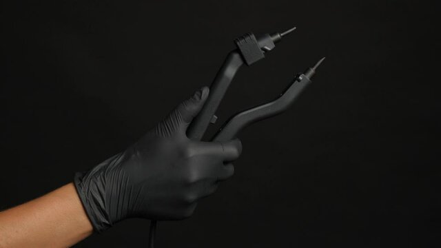 A hand with a black glove holds a hair extension tongs on a dark background