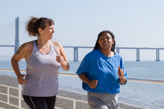 Happy plus size women doing sports near sea. Chubby women of different nationalities in sport clothes jogging at quay. Sport, body positive concept