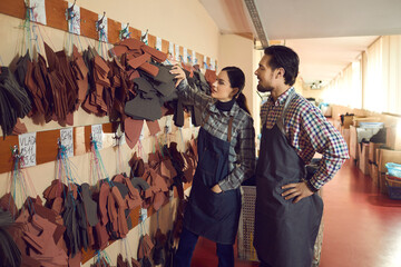 Shoe factory workers standing near wall of peg hangers with cut out natural leather details of...