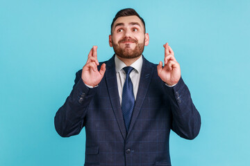 Portrait of hopeful man with beard wearing official style suit crossing fingers for luck, making a wish, dreaming of innermost, ritual. Indoor studio shot isolated on blue background.