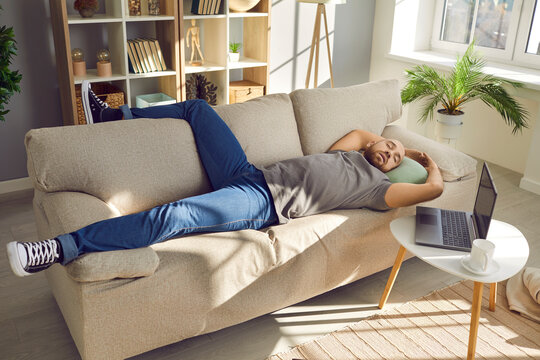 Lazy Man Enjoying A Nap. Tired Man Sleeping On The Sofa At Home. Young Guy Falls Asleep On The Couch While Working On His Laptop Computer In The Living Room