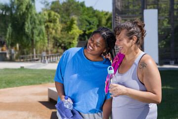 Merry plus size women resting after training. Chubby women of different nationalities in sport clothes drinking bottled water, laughing. Sport, body positive concept