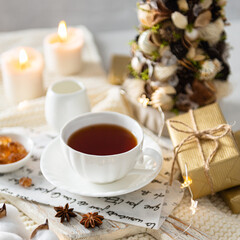Cozy winter composition with hot tea in a cup. Cup of black tea on wooden tray on knitted plaid with candles, presents and Christmas tree. Christmas menu, winter tea party, social media, square