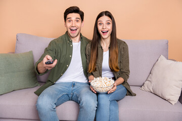 Photo of impressed spouses sit divan hold popcorn watch series remote control isolated over beige...