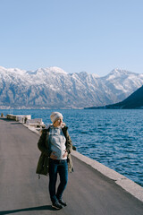 Pregnant woman in a jacket stands on the asphalt embankment and looks at the sea