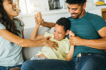 A happy family is spending their time together at home. The father is holding a boy and the mother is tickling him. - obrazy, fototapety, plakaty