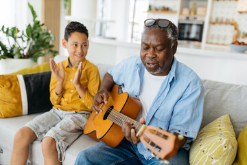 Musical African grandfather sitting at home with his grandson, playing the guitar and singing. His...