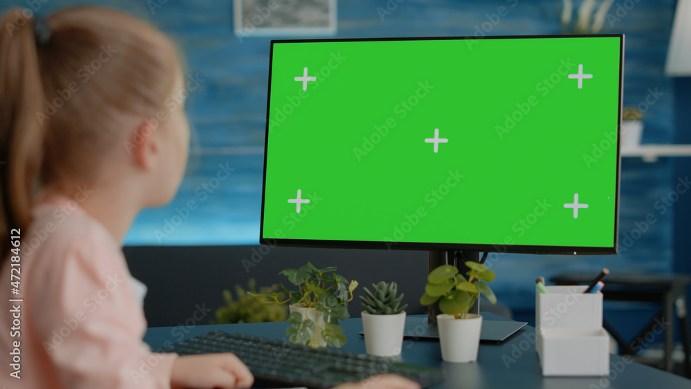 Poster young girl looking at monitor with horizontal green screen at desk. primary school child using compu