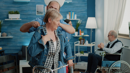 Disabled woman getting dressed with jacket while nurse giving assistance in nursing home. Medical assistant helping senior patient with walk frame for healthcare and chronic disease.