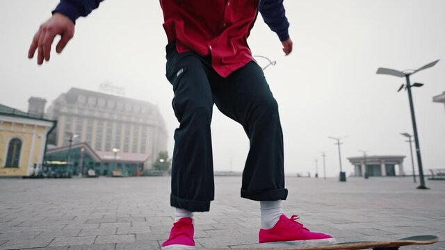 Young good looking skate boy in a purple jacket and red shoes is practicing longboarding at the skate in an empty city square. Skating youth hobby concept