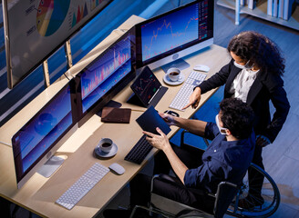 A stock investor who uses a wheelchair discussing with caucasian female colleague wearing face mask while she show thumb up in trading room with graph chart analysis screens and monitors
