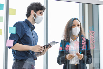Caucasian bearded businessman holding paper noting board and African American businesswoman in formal suit wearing face mask together while standing discussing consulting in conference meeting room