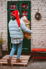 woman in a Santa hat carries a bag of gifts from the front door of the porch of the house.