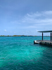 A turquoise water jetty in the Maldives overlooking an island with docked ships.