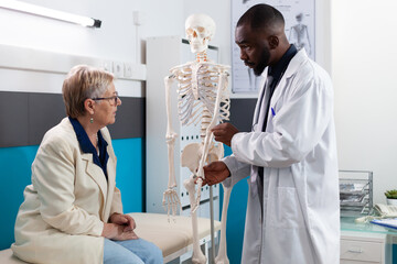 Specialist therapist doctor holding anatomical human skeleton arm explaining bones pain to senior retired woman patient during osteoporosis appointment in hospital office. Medicine concept