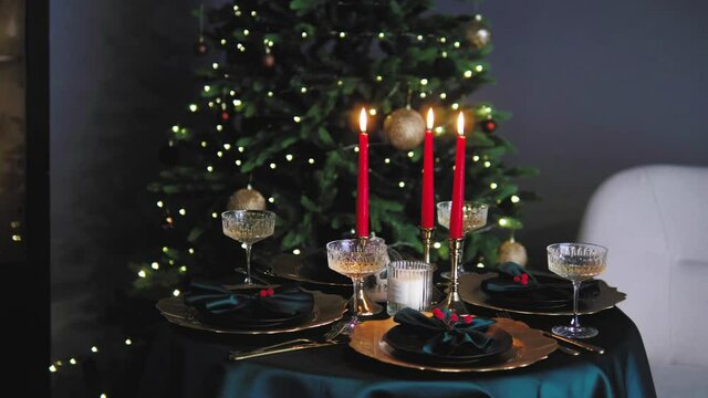Christmas Eve time celebration. Close up of prepaired decorated Christmas table in front of shiney Christmas tree with lights. Beautiful dishes: plates, wineglasses, candles, napkins. 