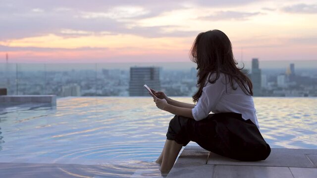 Asian woman resting is listening to music dancing alone moving to rhythm after work sitting by the edge of swimming pool and Blurred background skyscraper in city with Beautiful sunset or sunrise sky