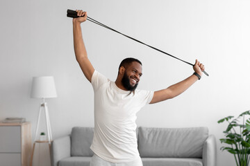 Smiling young african american bearded guy doing hand exercises with skipping rope in living room...