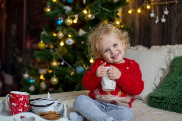 Cute toddler child, curly blond girl in a Christmas outfit, playing in a wooden cabin on Christmas, decoration around her. Child reading book and drinking tea