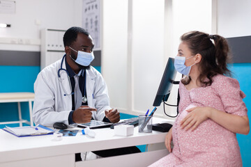 Fototapeta premium African american medic talking to pregnant woman at desk in medical office, wearing face masks at checkup appointment during pandemic. Specialist consulting adult with pregnancy.