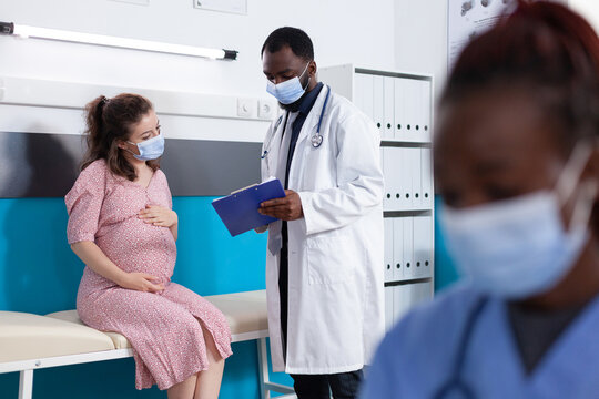 Physician showing checkup papers to pregnant woman in medical office. General practitioner doing examination with patient expecting child to check healthcare while wearing face masks - Powered by Adobe
