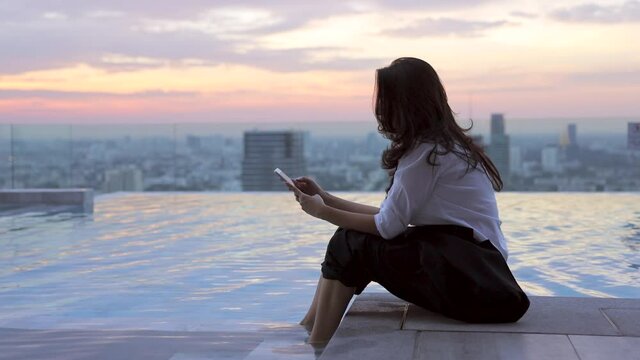 Easy relax asian businesswoman lifestyle after work at swimming pool with sunset and skyscraper in city view. Asian woman using mobile phone app playing game, shopping online, ordering delivery. 