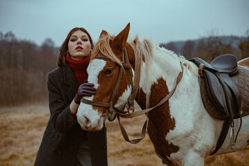 Fototapeta premium Elegant confident fashionable woman wearing classic grey woolen coat, turtleneck sweater, leather gloves posing with horse in nature. Outdoor autumn fashion portrait. Copy, empty space for text
