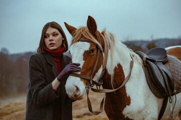 Obraz premium Outdoor autumn fashion portrait of elegant confident fashionable woman wearing classic grey woolen coat, turtleneck sweater, leather gloves posing with horse in nature. Copy, empty space for text
