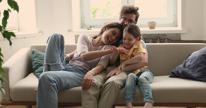 Family happiness. Portrait of young family couple with little preschool age daughter cuddle on soft couch at living room. Loving parents and small girl rest on sofa look at camera stack hands together