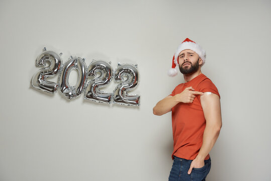 A serious man with a beard in a Santa hat is showing a shoulder with a plaster after vaccination against Coronavirus (Covid-19) near foiled silver balloons in the shape of 2022.