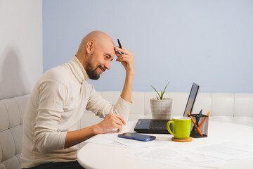 Thouthful bald man with beard in white turtleneck working on his notebook at home, side view