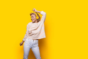 Fototapeta premium Joyful caucasian woman raises hand in dance move while listening music in headphones. Isolated over yellow bckground with copy space