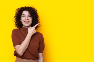 Front view of a happy woman standing isolated over yellow background looking at camera and pointing with index finger at emty space. Copyspace. Curly hair