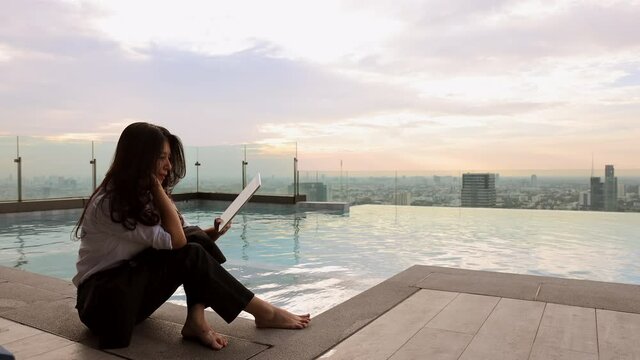 Freelancer asian woman resting after work with digital tablet outdoors at swimming pool with sunset and skyscraper view, enjoy her holiday or break