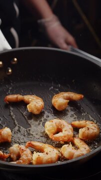 Professional chef roasts and tosses prawns in black frying pan with olive oil in restaurant kitchen. Tasty seafood cooking theme. Vertical video for smartphones, tablets in social media. Haute cuisine