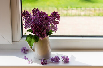 a purple bouquet of lilacs in a white jug stands on the windowsill of a country house.