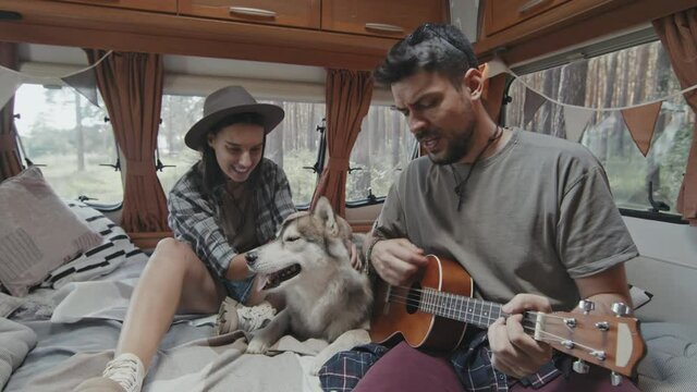 Slowmo medium shot of young man sitting on bed in cozy camper and playing song on ukulele. His happy girlfriend sitting beside him and singing while petting adorable husky