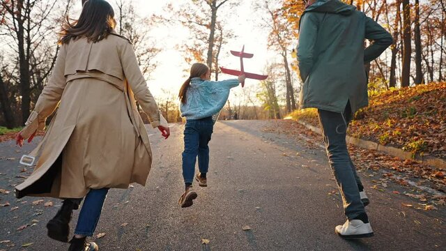 Young mother and father running with little daughter holding red toy plane. Autumn park. Close up, slow motion. Tracking dolly shot, back view
